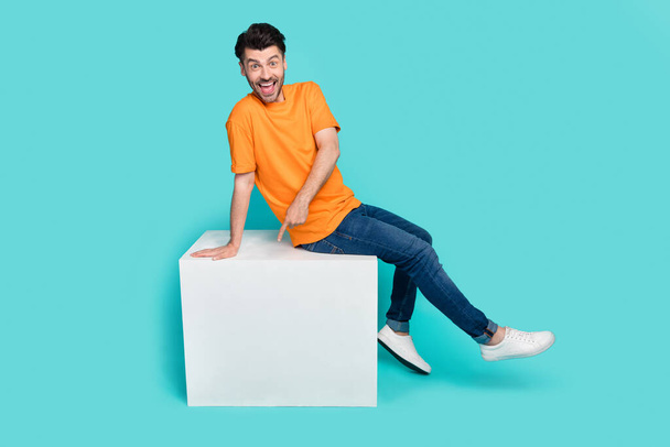 Full length photo of young funny attractive handsome toothy guy wear stylish outfit sitting white box finger pointing new information isolated on cyan color background. - Фото, изображение
