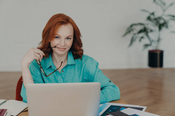 Beautiful happy busy female freelance worker with ginger hair, works online, uses laptop computer, prepares financial report at home, uses videoconferencing app, talks distantly with colleagues - Photo, Image