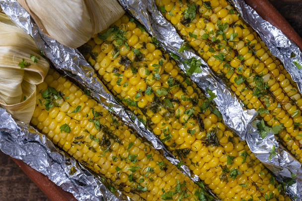 Tasty corn baked in foil with butter, garlic, chili, parsley and sea salt. Grilled corn in aluminum foil on brown wooden table. Top view, close up. Healthy food concept - Foto, afbeelding