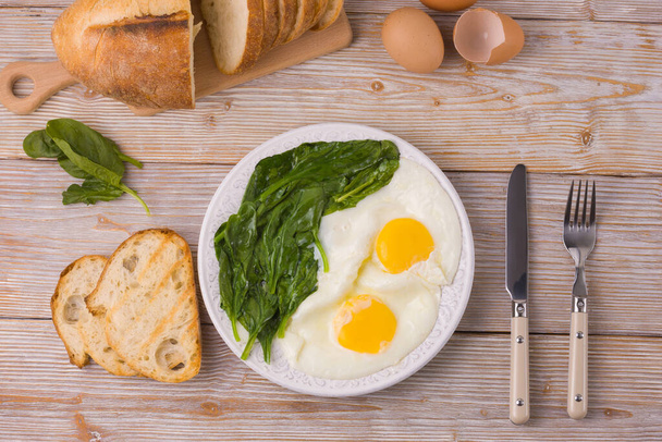 Plate with fried eggs and spinach on a light wooden background, table knife and fork, bread and croutons. View from the top. Breakfast on the table with fried eggs and spinach. - Photo, Image