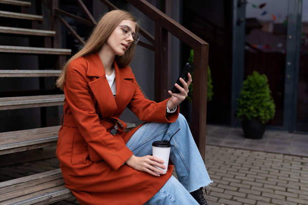 mladá stylová žena sedí na schodech vedle oblíbené kavárny s šálkem kávy a sleduje mobilní telefon. - Fotografie, Obrázek