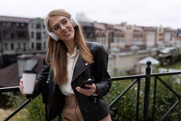 retrato de una joven alegre y feliz con un teléfono móvil y una taza de café en las manos escuchando música a través de auriculares con una sonrisa en la cara. - Foto, imagen