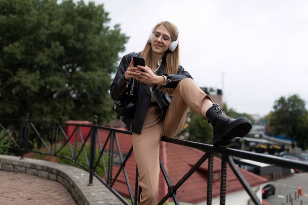 young woman listens to music in headphones sitting on the parapet on the background of the urban landscape. - Photo, Image