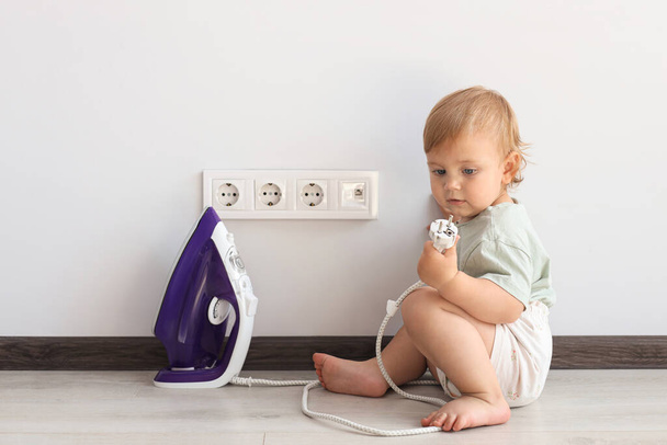 Cute baby playing with electrical socket and iron plug at home. Dangerous situation - Фото, зображення