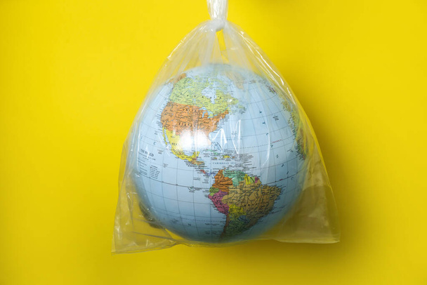 Globe in plastic bag hanging against yellow background. Environmental conservation - Foto, imagen