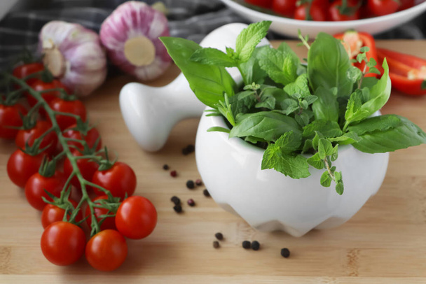 Mortar with different fresh herbs near pepper, garlic and cherry tomatoes on wooden table, closeup - Fotografie, Obrázek
