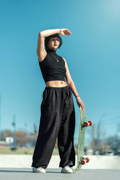 Menina skatista muito legal com óculos de olho posando com sua prancha de skate em direção à luz solar. Fotografia vertical - Foto, Imagem