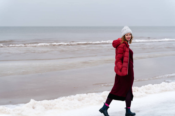 A woman in a red jacket and a red knitted dress walks in winter along the sea. - Foto, Imagen