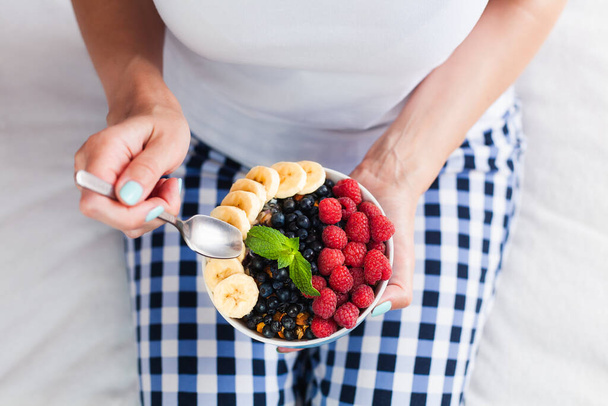 The girl eats a healthy breakfast of muesli with fresh fruits and berries while sitting on the bed at home. Healthy food concept. ?lose-up. Top view. - Photo, Image