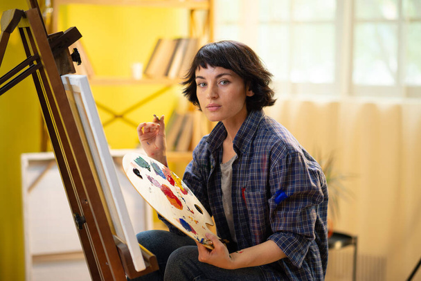 A beautiful woman wearing a blue flannel t shirt is painting something amazing and modern on her canvas in a big and brightly lit art atelier. Portrait - Foto, imagen