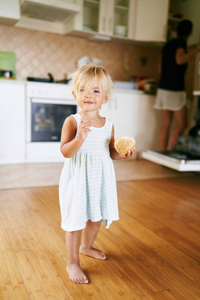 Ein kleines Mädchen mit einem Brötchen in der Hand steht auf einem Holzboden in der Küche. Hochwertiges Foto - Foto, Bild