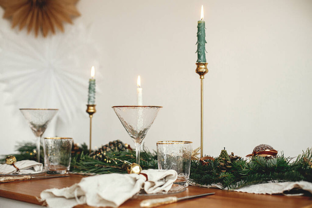 Стильная сервировка рождественского стола. Белье салфетка с колокольчиком на тарелке, винтажные столовые приборы, винное стекло, ель ветви с золотыми огнями, сосновые шишки и свечи на столе. Атмосферный праздничный бранч - Фото, изображение