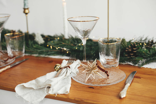 Christmas table setting. Linen napkin with bell, star on plate, vintage cutlery, glasses, fir branches with golden lights and pine cones on table. Holiday arrangement of table - Foto, Bild
