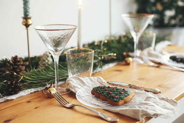 スタイリッシュなクリスマステーブルの設定。プレート上のクリスマスツリークッキー、鐘とリネンナプキン、ヴィンテージカトラリー、ワイングラス、金色のライトを持つモミの枝、テーブルの上のキャンドル。大気の休日ブランチ - 写真・画像