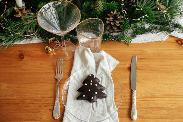Christmas table setting. Christmas tree cookie on plate, linen napkin with bell, vintage cutlery, glasses, fir branches with golden lights and pine cones on table. Modern Holiday arrangement - Photo, Image