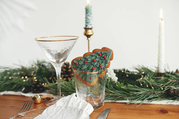 Stilvolle weihnachtliche Tischdekoration. Weihnachtsbaum-Plätzchen im Weinglas, Teller mit Leinenserviette, Vintage-Besteck, Tannenzweige mit goldenen Lichtern, Kerzen auf dem Tisch. Weihnachtsessen, kreativer Tisch gedeckt - Foto, Bild