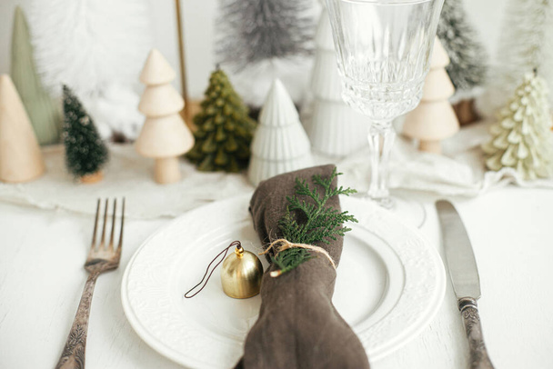 Mesa de Navidad. Elegante servilleta con campana y abeto en el plato, cubiertos vintage, vasos, pequeños árboles de Navidad festivos en la mesa rústica blanca. Arreglo de vacaciones de mesa - Foto, imagen