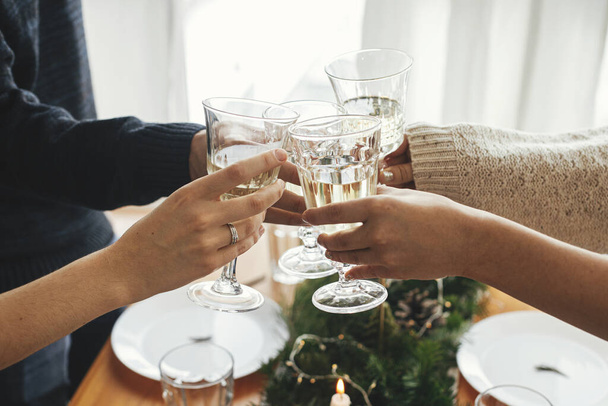 Hands with champagne glasses clicking on background of stylish table setting with fir branches with golden lights and candles. Friends toasting with champagne and celebrating. Christmas feast - Foto, Bild