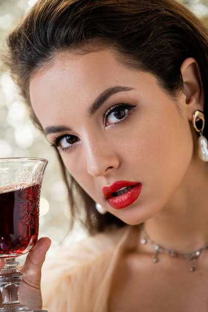 πορτρέτο της νεαρής γυναίκας με μακιγιάζ κοιτάζοντας κάμερα κοντά σε ποτήρι κόκκινο κρασί σε λευκό φόντο - Φωτογραφία, εικόνα