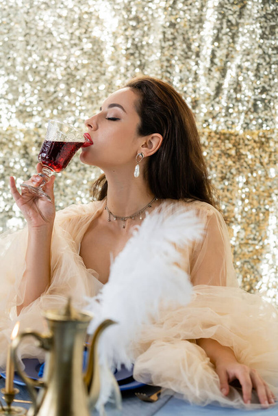 glamour woman in tulle dress drinking red wine near blurred copper jug on glitter background - Photo, Image