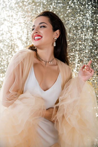 happy brunette woman in elegant dress and makeup looking away on shiny silver background - Fotó, kép