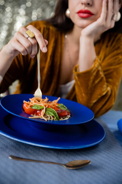 vista recortada de la mujer borrosa sosteniendo tenedor cerca de placa azul con espaguetis sobre fondo brillante - Foto, Imagen