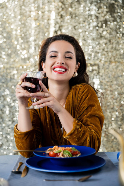 radosna i elegancka kobieta ze szklanką czerwonego wina patrząca na kamerę w pobliżu spaghetti na błyszczącym srebrnym tle - Zdjęcie, obraz