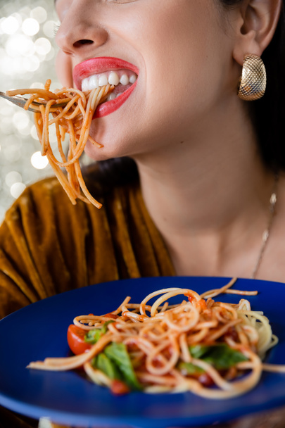 partial view of woman with red lips and golden earring eating pasta from blue plate on shiny background - Photo, Image