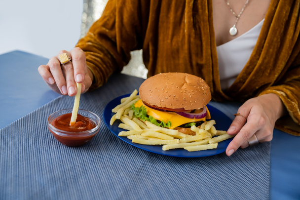 oříznutý pohled rozmazané ženy namáčení hranolky do kečupu v blízkosti talíře s chutným burger na šedém pozadí - Fotografie, Obrázek