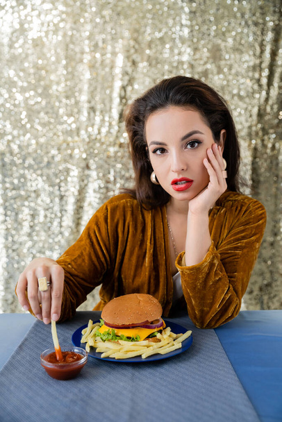 mulher morena em veludo vestido segurando batatas fritas perto de ketchup e hambúrguer enquanto olha para a câmera no fundo brilho - Foto, Imagem