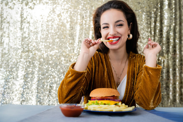 stylish and cheerful woman looking at camera and eating french fries near burger on shiny silver background - Foto, Bild
