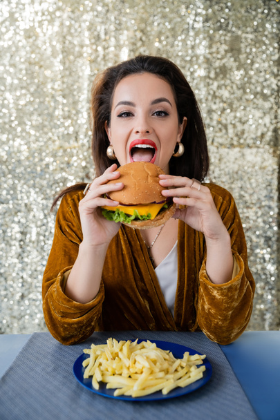morena mujer elegante mirando a la cámara mientras come hamburguesa cerca de papas fritas sobre fondo plateado brillante - Foto, Imagen