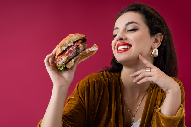 happy and trendy brunette woman holding delicious burger isolated on red - Photo, Image
