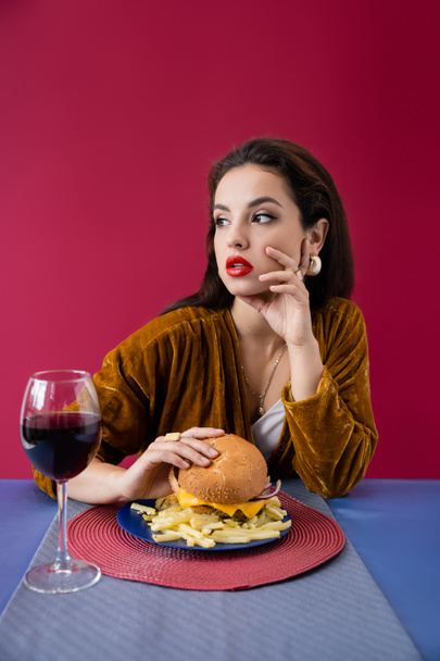 stijlvolle vrouw met hamburger weg te kijken in de buurt van frietjes en wijnglas geïsoleerd op rood - Foto, afbeelding
