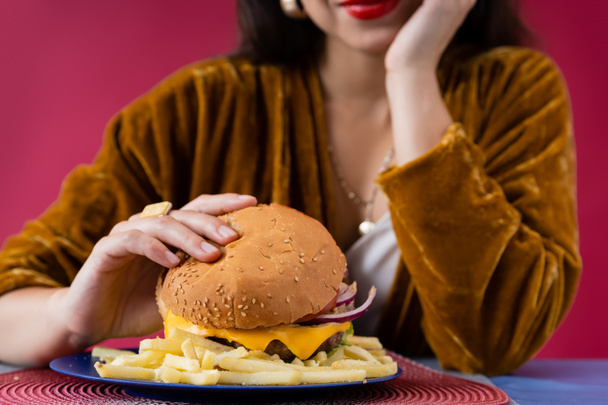 vista recortada de mujer borrosa en vestido de terciopelo sosteniendo hamburguesa cerca de papas fritas aisladas en rojo - Foto, Imagen