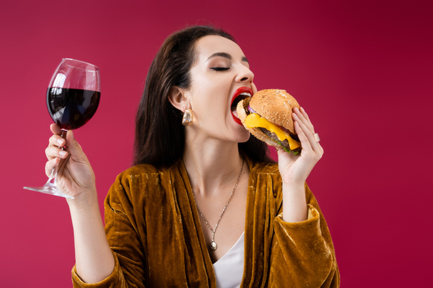μελαχρινή γυναίκα σε βελούδινο φόρεμα κρατώντας ένα ποτήρι κρασί και τρώγοντας νόστιμο burger απομονώνονται σε κόκκινο - Φωτογραφία, εικόνα
