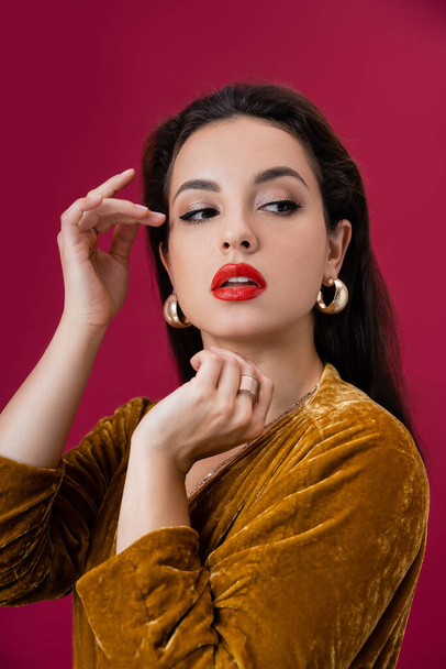 glamour woman in golden earrings and velour dress holding hands near face and looking away isolated on red - Foto, Imagen