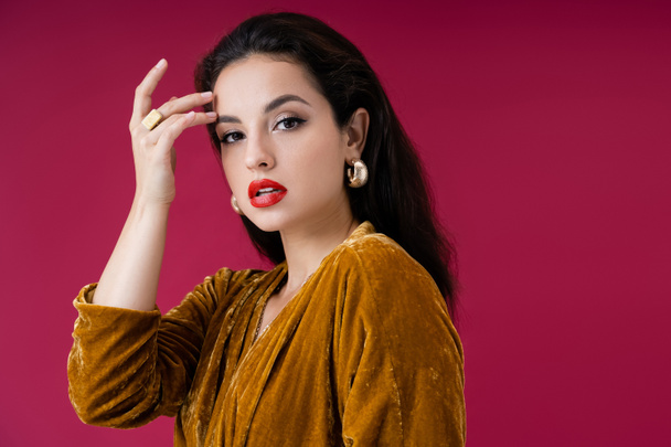 charming woman in velour dress and golden earrings touching face and looking at camera isolated on red - Foto, immagini
