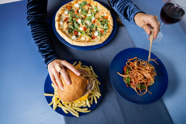 partial view of woman taking burger and pasta near french fries and pizza served on blue table - Photo, Image