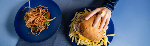 top view of cropped woman taking burger near french fries and spaghetti on blue plates, banner - Photo, Image