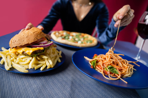 ピザの近くにフライドポテトとスパゲティのハンバーガーと赤で隔絶された背景にぼやけた女性の選択的な焦点 - 写真・画像