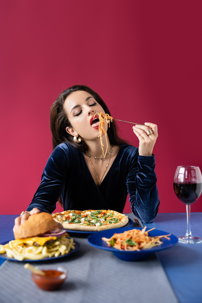 elegant woman with closed eyes eating delicious pasta near wine and meal served on blue table isolated on red - Photo, Image