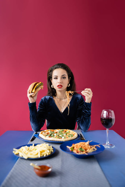 mujer elegante con hamburguesa y espaguetis cerca de la mesa azul con papas fritas y copa de vino aislado en rojo - Foto, Imagen