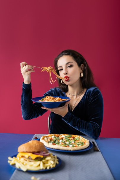 stylish woman in velour dress eating spaghetti near burger with french fries and pizza isolated on red - Zdjęcie, obraz
