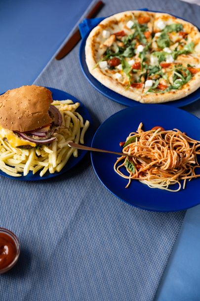 πάνω άποψη των σπαγγέτι κοντά τηγανητές πατάτες με burger και θολή πίτσα σε μπλε πιάτα - Φωτογραφία, εικόνα