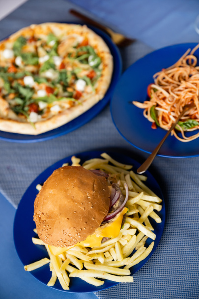 Teller mit verschwommener Pizza in der Nähe von Spaghetti und Pommes mit Burger auf blauem Tisch - Foto, Bild