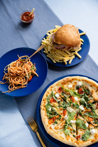 vrchní pohled na hranolky s hamburgerem v blízkosti chutných těstovin a pizzy na modrých talířích - Fotografie, Obrázek