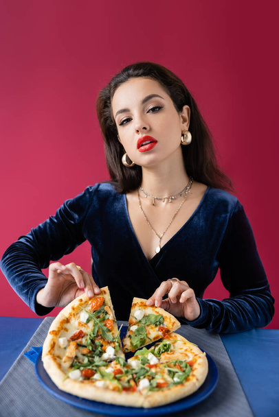 sensual woman in blue velour dress and golden accessories taking piece of delicious pizza isolated on red - Photo, Image