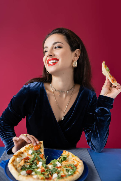 glückliche Frau in blauem Velourkleid, die ein Stück Pizza in der Hand hält und isoliert auf Rot wegschaut - Foto, Bild