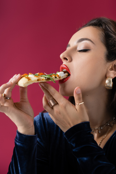 κομψή και σέξι γυναίκα σε χρυσά δαχτυλίδια δάχτυλο τρώει νόστιμη πίτσα απομονωμένη σε κόκκινο - Φωτογραφία, εικόνα